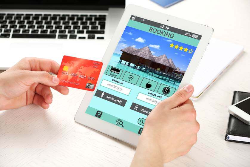 A person holds a tablet displaying a hotel booking app, while holding a credit card in the other hand, with a laptop in the background.