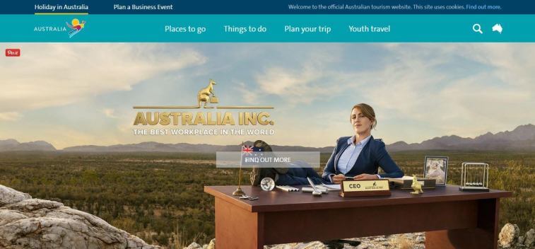 A woman in a business suit sitting behind an office desk placed outdoors in a scenic Australian landscape with mountains in the background, promoting Australia tourism.