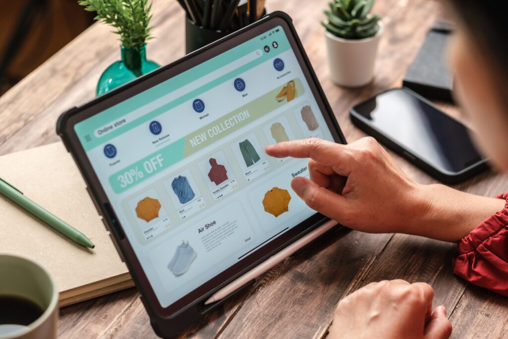 A close-up of a person's hand using a tablet to shop online, with the screen displaying a clothing store app.