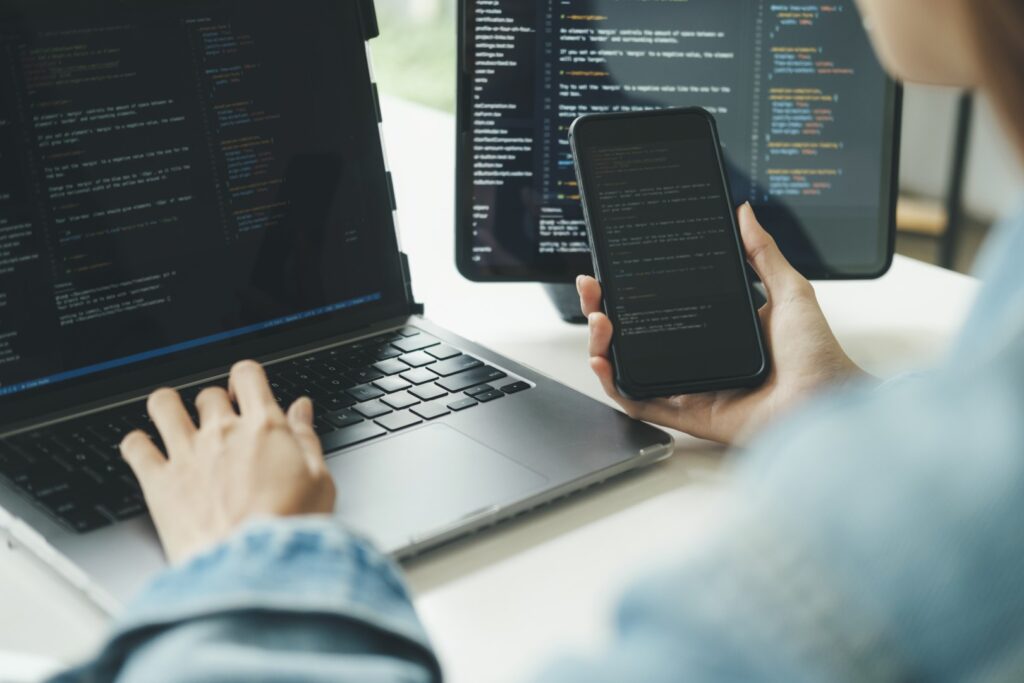 A programmer wearing a denim shirt types on a laptop while holding a smartphone displaying code, with multiple monitors showing code in the background.