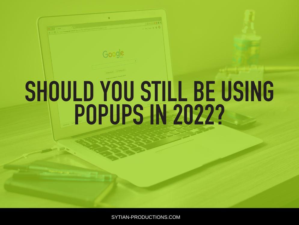 A laptop sits on a wooden desk, overlaid with a green tint and the text "SHOULD YOU STILL BE USING POPUPS IN 2022?".