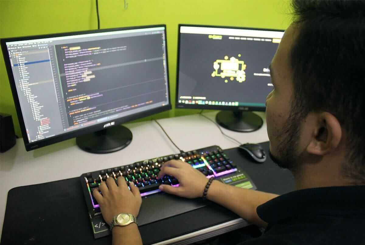 A young man sits in profile before two computer monitors, typing code on an RGB backlit keyboard at a desk against a lime green wall.