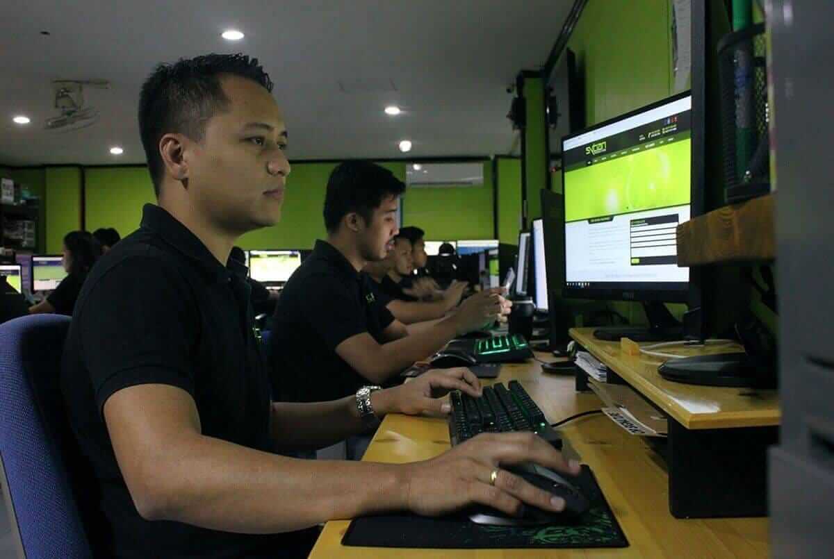 A side angle of young men sitting at wooden desks with computers in an office room, with green walls and black clothes.