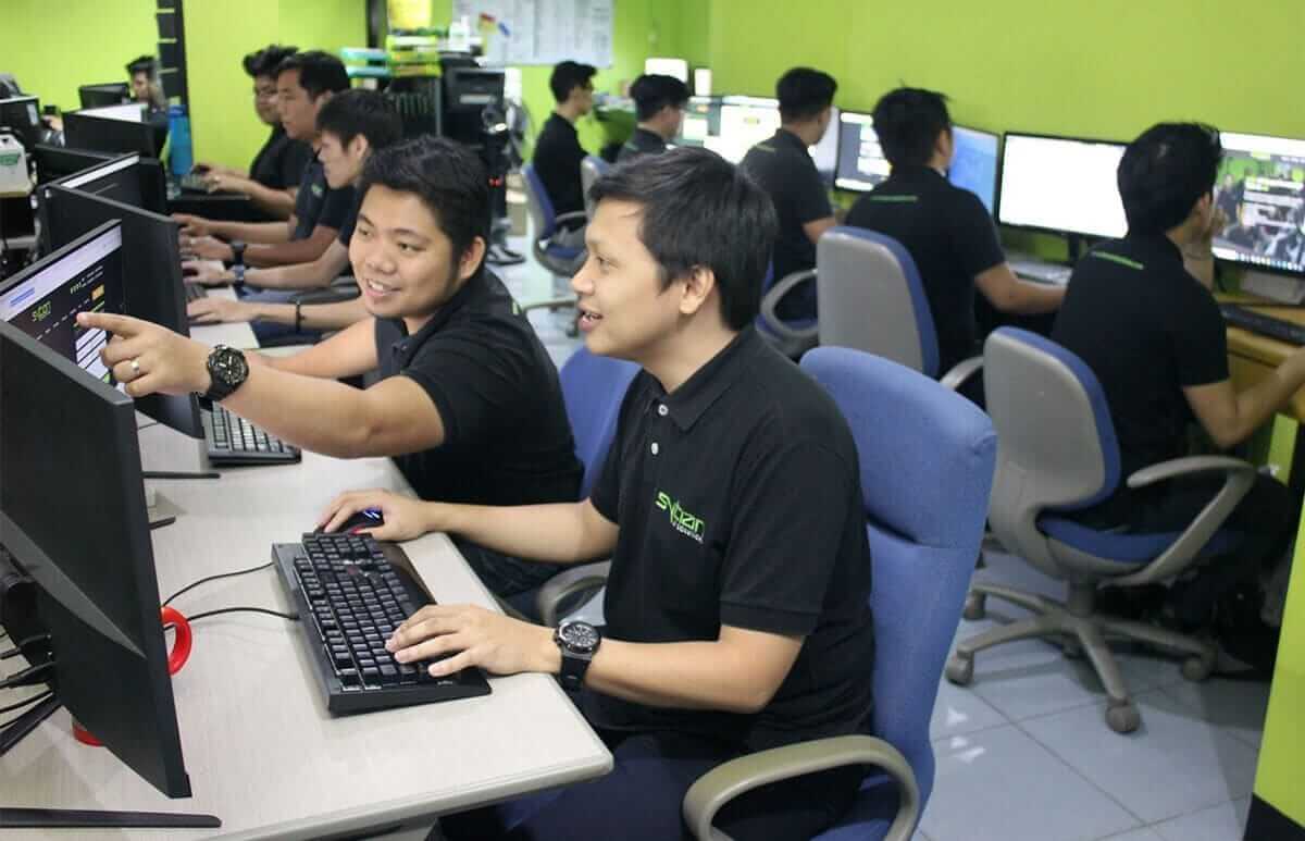 Several people are working at computers in an office with bright green walls, with two men in the foreground collaborating and looking at a monitor.