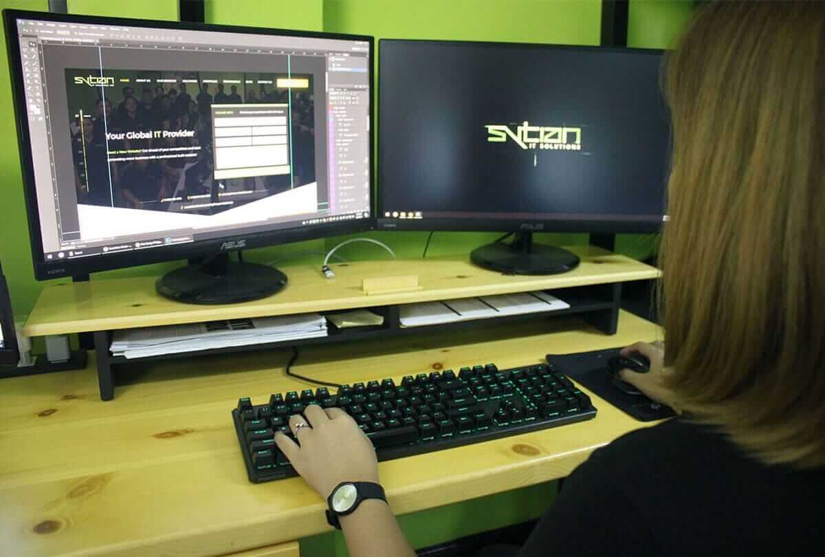 A woman with blonde hair sitting at a wooden desk with two computer monitors, typing on a black keyboard.