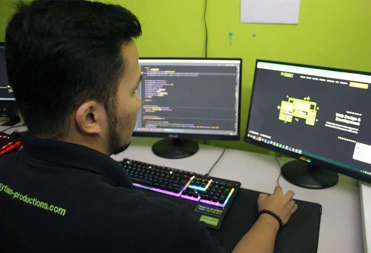 A web developer sits in front of a dual-monitor setup, viewing code on the left screen and a website preview on the right.