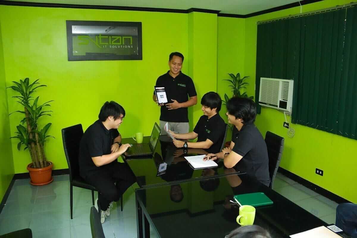 Four employees in black shirts gather around a black conference table in a bright green room with an IT solutions logo on the wall.