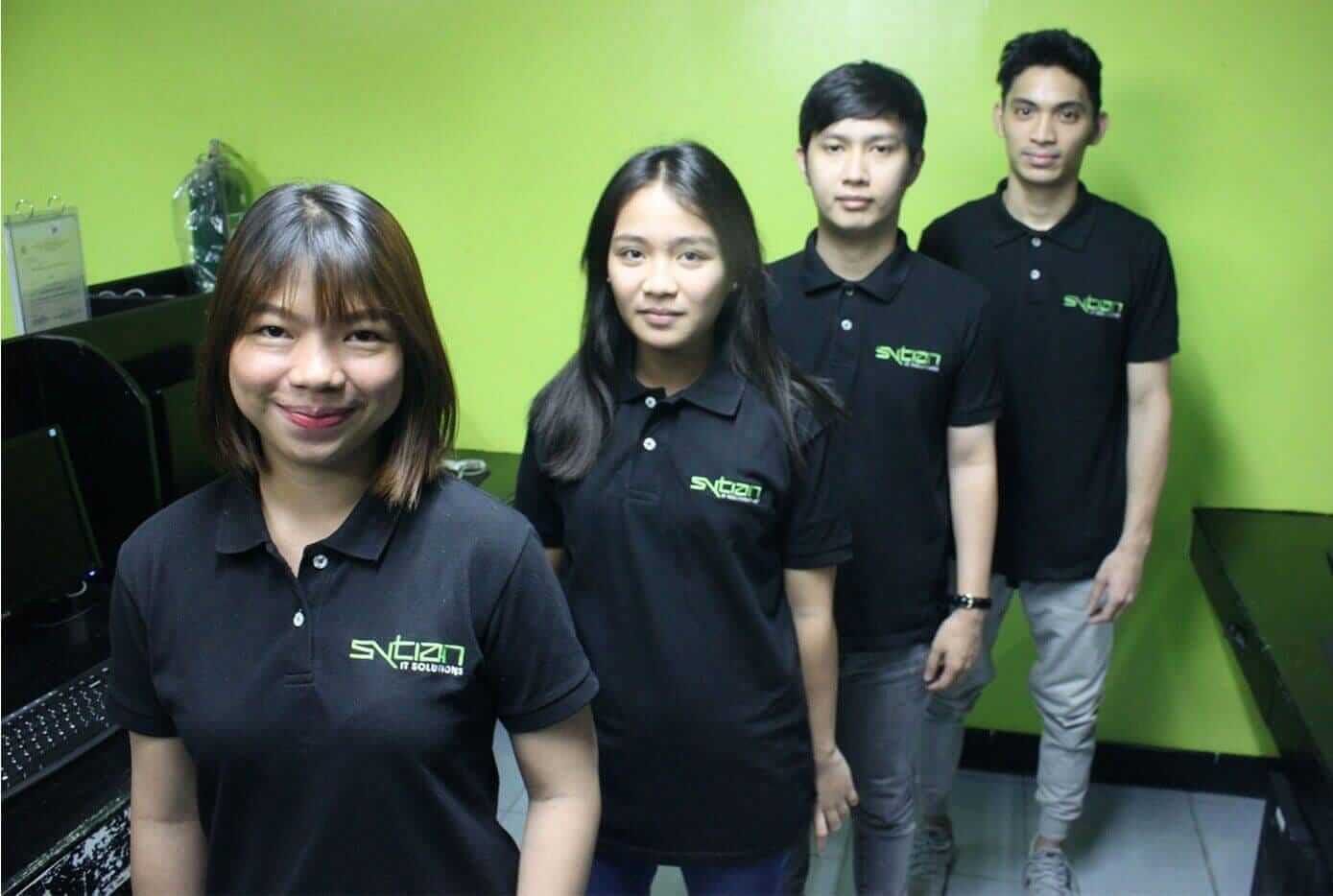 Four individuals, two women and two men, pose in front of a green wall, all wearing black polo shirts featuring a white company logo on the chest.