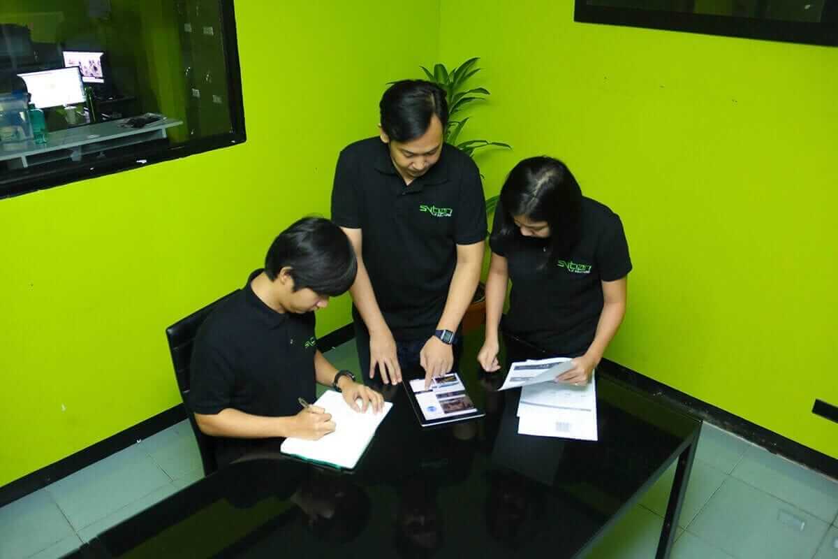 Three individuals in black polo shirts review documents together in an office with bright green walls.