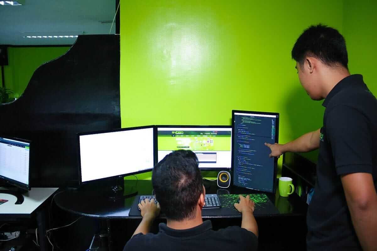 A man sits at a desk with three computer monitors, and another man stands beside him, pointing at the screen on the right.