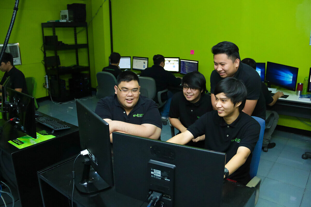 Group of young male programmers in matching black polo shirts smiling and working collaboratively on computers in an office with bright green walls.